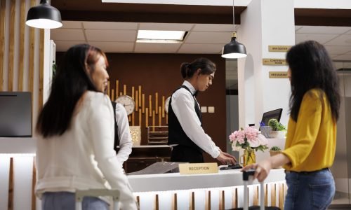 Cozy hotel reception with two female guests waiting for concierge to finalize booking procedure. Relaxed customers holding baggages getting ready to check in at lavish resort