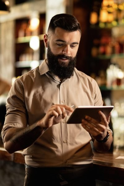 Smiling barista using digital tablet while working in a bar. Young smiling waiter using touchpad while working in a pub.